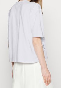 Camiseta blanca de algodón con mangas cortas, cuello redondo y un corte relajado. Presenta un detalle decorativo a lo largo de la costura lateral.