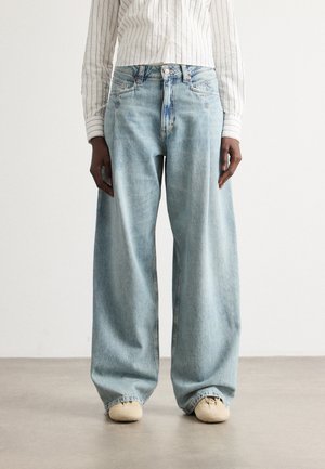 Persona che indossa jeans in denim azzurro a gamba larga, scarpe beige e una camicia bianca con bottoni a righe verticali sottili, in piedi su un pavimento grigio.
