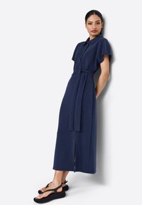 Abito midi blu navy con maniche corte, apertura con bottoni, cintura in vita e spacco frontale. Indossato con sandali neri con suola piatta.