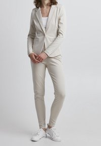 Blazer à carreaux beige et pantalon assorti, tous deux en tissu lisse. Portés avec des baskets blanches. Présente une coupe ajustée et des revers crantés.