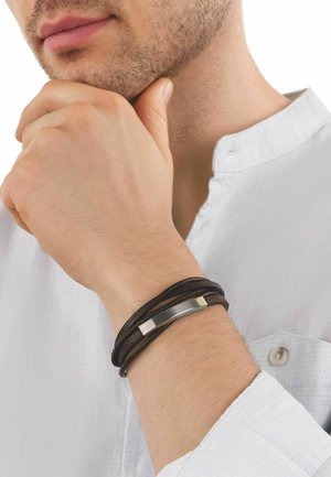 Mann trägt weißes Hemd, die Hand am Kinn, zeigt ein braunes Lederwickelarmband mit einer silbernen Metallplatte am Handgelenk.