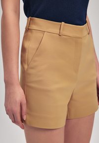 Brune skræddersyede shorts med en glat tekstur, der har bæltestropper og sidelommer. Båret med en mørkeblå strikket top.