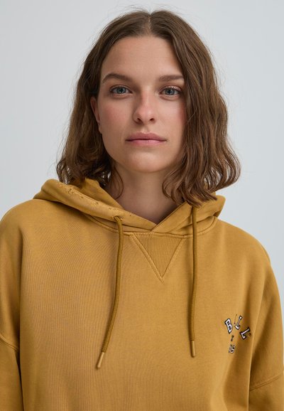 Dzeltena sinepju krāsa kapuce ar priekšējo kabatu, aukliņu kapuci un nelielu logo uz krūšu. Mīksta tekstūra un brīvs griezums.