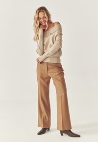 Beżowy sweter typu off-shoulder z detalami w postaci guzików, w połączeniu z brązowymi spodniami typu wide-leg i ciemnymi butami na obcasie na jasnym tle.