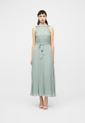 Kvinde stående iført ærmeløs salviegrøn blonderkjole med plisseret nederdel og bundet taljebælte, kombineret med beige ankelrem-sandaler.