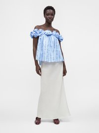Femme portant un haut bleu décolleté aux épaules avec de grandes fleurs en tissu et une longue jupe blanche, debout devant un fond uni.