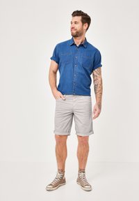 Men's short-sleeve denim shirt in dark blue with two front pockets, paired with light gray rolled-up shorts and brown sneakers.