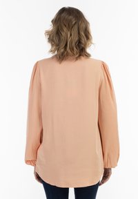 Ljus persikofärgad blus med långa ärmar, tillverkad av ett mjukt tyg. Har volangärmar och en avslappnad passform, med en rundad fåll.