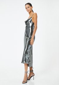 Metallisch graues Midi-Kleid mit Spaghettiträgern, drapiertem Ausschnitt und glatter Textur. Kombiniert mit schwarzen Riemchenheels, die funkelnde Akzente aufweisen.