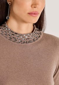 Vrouw in een bruine gebreide trui met zwarte ovale kralen op de kraag en een lange gouden oorbel met twee gekleurde stenen.