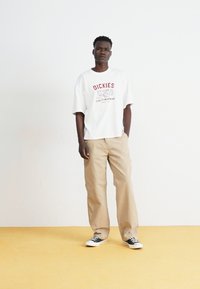 Bela oversize majica s rdečim logotipom "Dickies", bež široke hlače in črno-bele superge na rumenem tleh.