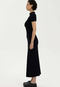 Czarna dopasowana sukienka z krótkimi rękawami, wysokim dekoltem i długą sylwetką. Gładka tekstura bez wzorów. Noszona z czarnymi klapkami.