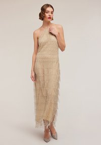 Beaded halter neck dress in beige with tiered fringe detailing. The silhouette is form-fitting, paired with sparkly pointed-toe heels.
