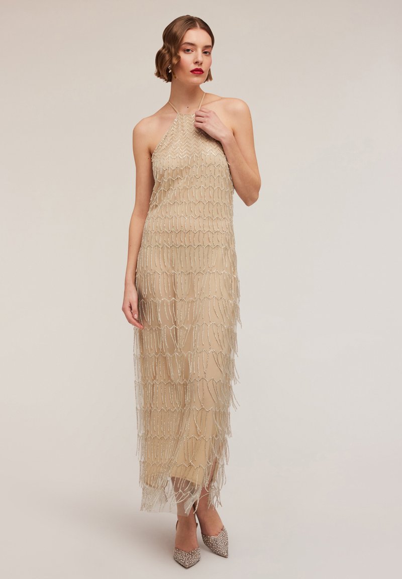 Beaded halterneck dress in beige with tiered fringe detailing. The silhouette is form-fitting, paired with sparkly pointed-toe heels.
