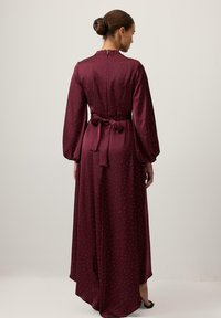 Barna hajú nő kontyba fogva, hosszú ujjú burgundi színű ruhát visel, amelyen apró fehér pöttyök és egy megkötött derékmasni található, egy üres fal előtt áll.