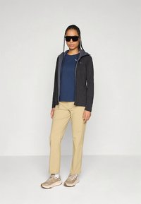 Casaco leve preto com capuz, t-shirt azul-marinho e calças bege. Modelo usa óculos de sol e ténis desportivos bege. Textura de tecido lisa.