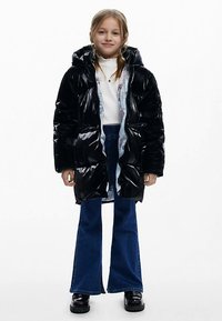 Zwarte glanzende pufferjas met capuchon, voorzien van een blauw bloemenprint voering, gecombineerd met een witte trui, gebloemde blauwe jeans met uitlopende pijpen en zwarte plateau schoenen.