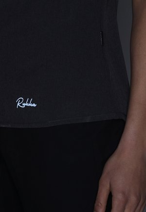 Primer plano de una persona que lleva una camiseta negra con el logo blanco "Rukka" cerca del borde y una mano derecha visible apoyada al lado.