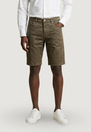 Persona de piel oscura que lleva pantalones cortos verde oliva hasta la rodilla, camisa blanca de manga larga y zapatillas blancas, de pie con una mano en el bolsillo.