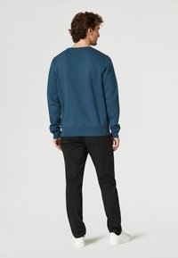 Sudadera de algodón azul con cuello redondo, puños y dobladillo acanalados, combinada con pantalones negros y zapatillas blancas, vista desde la parte de atrás.