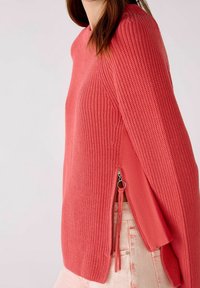 Pull en tricot corail avec des manches longues et des fermetures éclair sur les côtés. Présente un motif texturé et une coupe décontractée. Accentué par un détail à cordon de serrage.