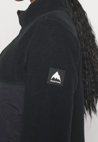 Czarna bluza polarowa z wysokim kołnierzem, z gładkim przednim panelem i kwadratową naszywką z białym logo góry na lewym ramieniu.