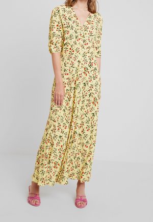 Kvinde iført en lang gul kjole med rød og grøn blomsterprint og lyserøde sandaler med åben tå, stående mod en ensfarvet lys baggrund.