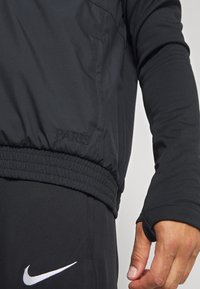 Sort sportsjakke med elastisk talje og manchetter, med "PARIS" broderet på siden, parret med sorte bukser og et hvidt Nike-logo.