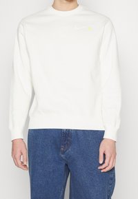 Sudadera blanca con cuello redondo, puños y dobladillo acanalados. Presenta un logo de Nike en azul claro y un pequeño detalle de corazón amarillo.