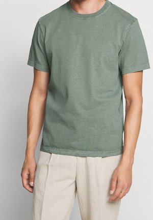 Mężczyzna ubrany w zieloną, prostą, krótką koszulkę z okrągłym dekoltem i jasnobeżowe spodnie, stojący na neutralnym tle.