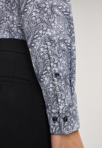 Camicia a maniche lunghe con motivo paisley in bianco e nero. Presenta bottoni scuri sui polsini e un indumento aderente nero sotto.