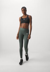 Zwarte sport-bh met wit logo, gecombineerd met groene leggings met zijstreepjes. Model draagt zwarte sportschoenen.