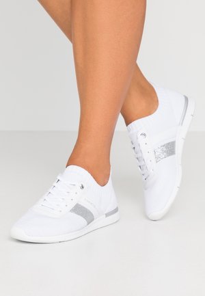 Persoon draagt witte sneakers met een zilveren glinstertreep en een elastische enkelkraag, staand op een lichtgrijze vloer.