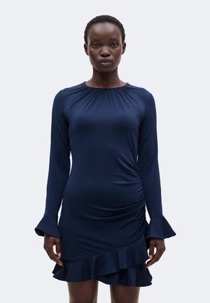 Femme aux cheveux courts portant une robe bleu marine à manches longues avec des poignets à volants et un ourlet à volants asymétrique, debout devant un fond uni.