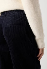 Close-up van een persoon in donkerblauwe corduroy broek en een crèmekleurige fuzzy trui met de hand rustend op het bovenbeen.