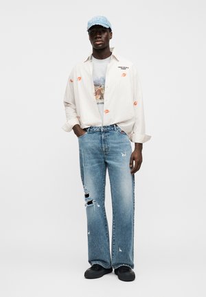 Langärmliges, leicht gestreiftes Hemd mit orangen Lippenabdrücken, graues grafisches T-Shirt, zerrissene blaue Jeans und eine verblasste blaue Kappe. Schwarze Schuhe.