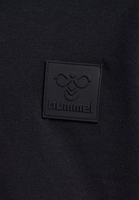 Svart stickad tyg med en strukturerad logopatch i svart gummi med märkesnamnet "hummel" i centrum.