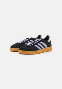 Black suede sneakers feature three horizontal white stripes, a rubber gum sole, and white laces, with a slight heel and a rounded toe.