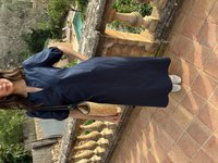 Žena stoji na terakota pločicama kraj kamenog balustrada, nosi tamnoplavu haljinu, bijele tenisice bez vezica, drži pletenu slamnu torbu.