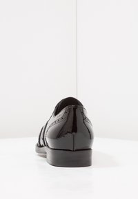 Chaussure de ville en cuir verni noir avec des détails brogue, vue de l'arrière, sur une surface blanche et réfléchissante.
