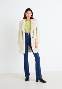 Krämfärgad fuskpälsjacka med bred krage och dubbelknäppta knappar, i kombination med högmidjade jeans och svarta klackiga ankelstövlar.