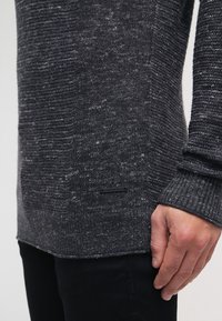 Pull en tricot gris avec un motif texturé, des poignets côtelés et des détails sur les côtés. Comprend une petite étiquette de marque noire en bas.