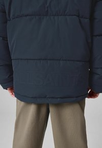 Jūras zila pūkainā jakas ar quilted dizainu un reljefu "ALLSAINTS" logo apakšmalē, apvienota ar gaiši olīvkrāsas biksēm.