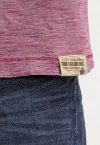 T-paita, jossa on tummanpunaiset raidat ja kudottu etiketti ommeltuna helmaan, yhdistettynä tummiin farkkuihin, joissa on hienovarainen rakenne.