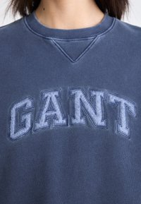 Námořnická modrá mikina se strukturovaným logem "GANT" ve světle šedé barvě. Hladký límec a měkká, mírně kartáčovaná textilie.