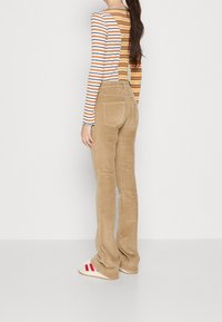 Femme portant un pantalon en velours côtelé beige, un haut à manches longues rayé à double motif, et des baskets blanches avec des rayures rouges, se tenant contre un mur uni.