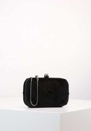Petit sac pochette noir matelassé avec une chaîne argentée et un fermoir rond, posé sur un piédestal blanc sur un fond uni.
