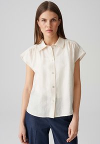 Weißes Kurzarm-Hemd mit Knöpfen und Kragen, aus leichtem Stoff, mit einer leicht zerknitterten Textur und einem abgerundeten Saum.