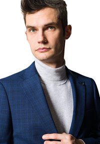 Niebieska marynarka w kratę z wyraźnymi klapami, noszona na szarym golfie. Gładka tekstura, dopasowany krój oraz detal w postaci przypinki na marynarce.