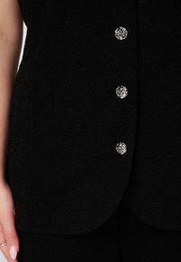 Vêtement noir texturé avec un ourlet courbé et cinq boutons argentés brillants sur le devant, présentant un détail de surface à motifs.